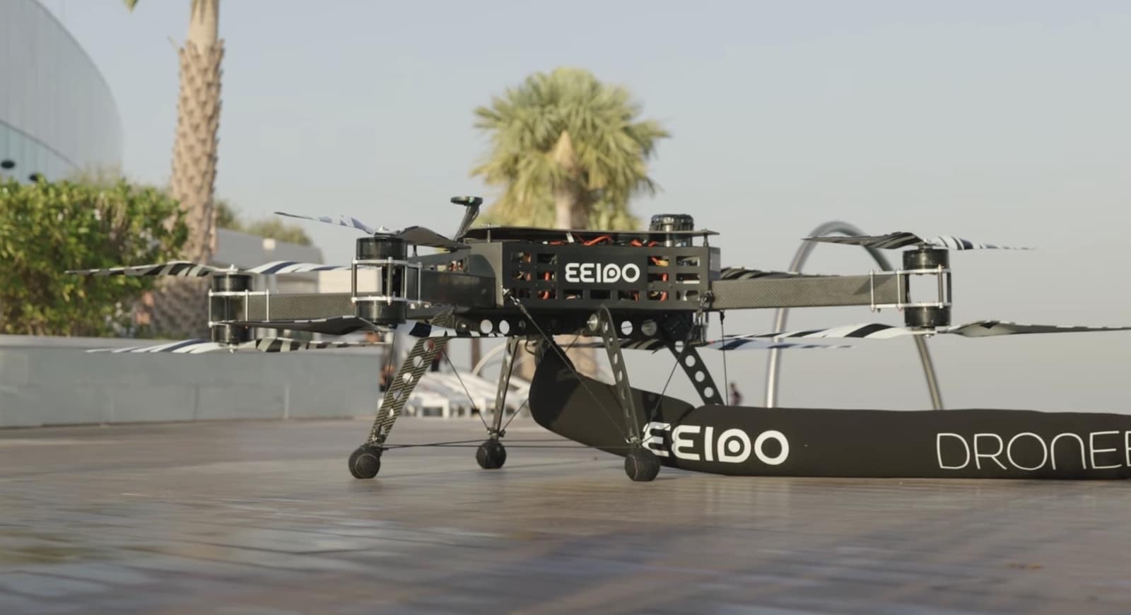 A large heavy-lift multirotor drone standing on a rooftop pad among palm trees.