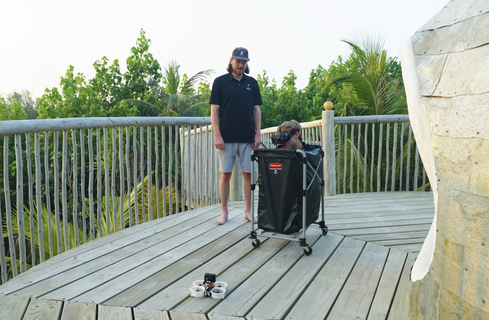 FPV pilot stands on a resort deck beside a gear cart and drone.
