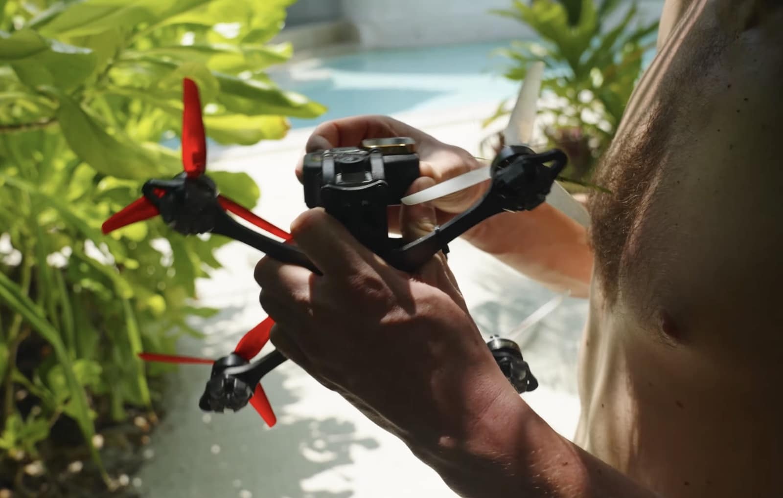 Hands hold a cinewhoop FPV drone with red propellers beside a pool.