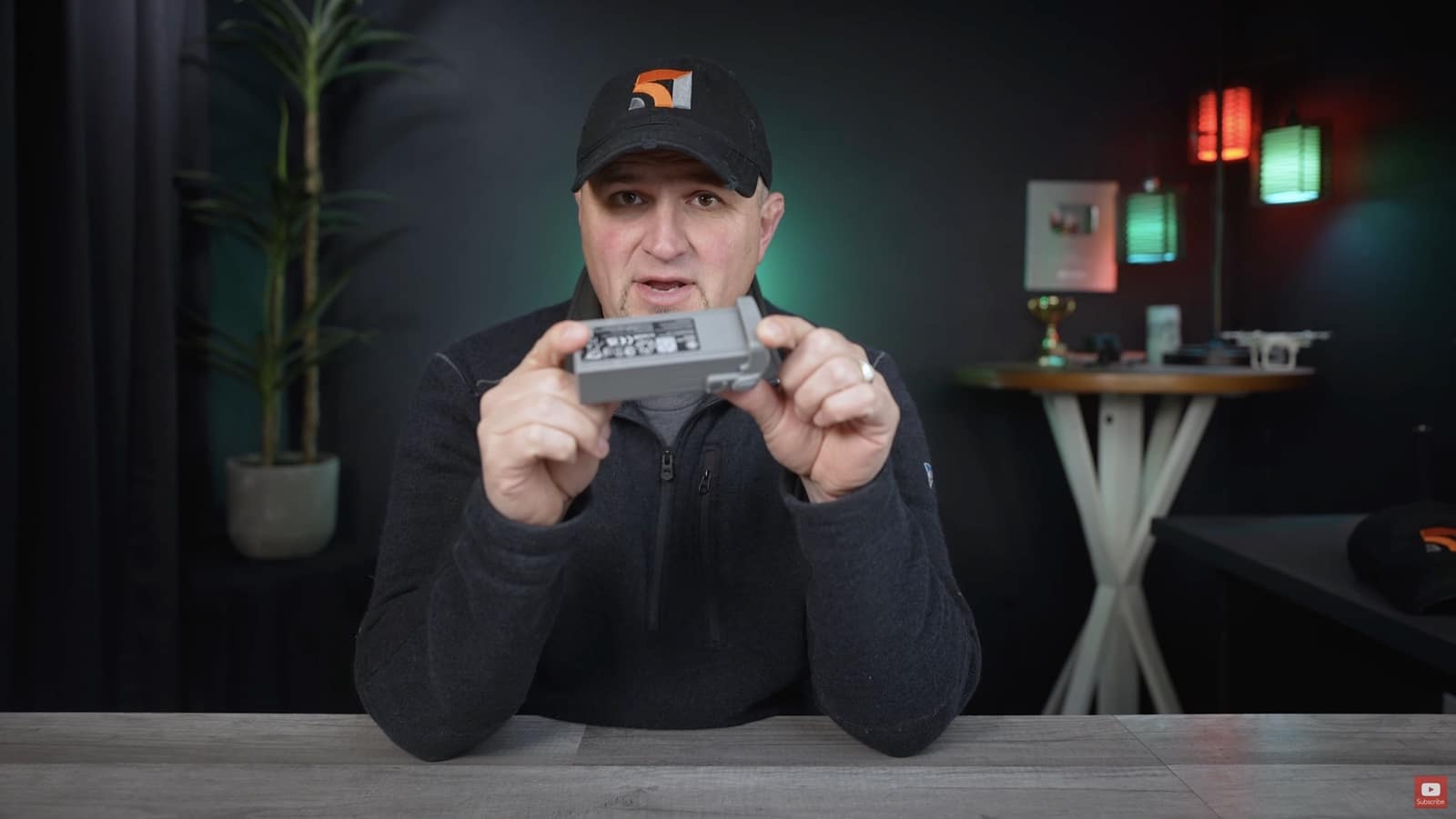 Presenter holds a drone battery in both hands at a studio desk.