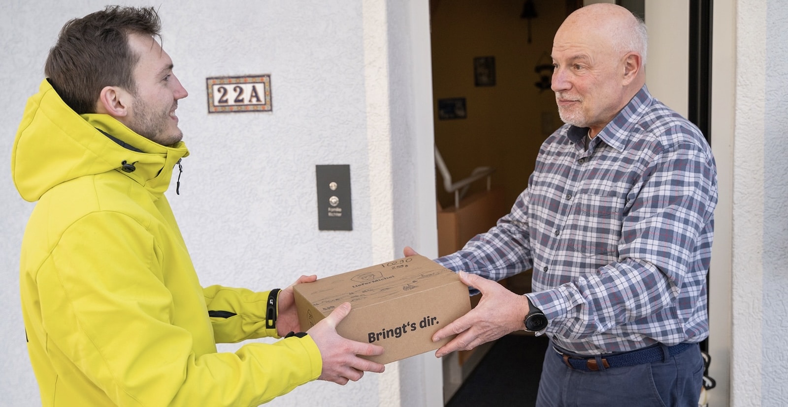 Courier in a yellow jacket hands a cardboard parcel to a man at his door.