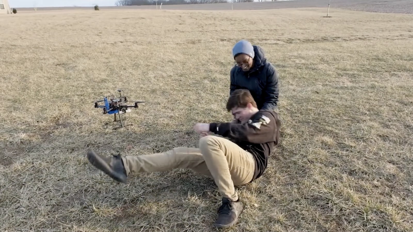 Bystander tends to a collapsed volunteer beside a drone landed in a field.