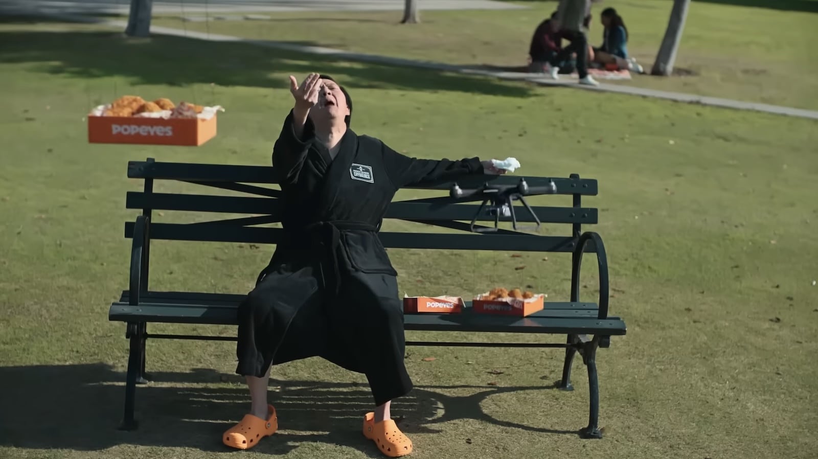 Ken Jeong on a park bench as a drone delivers Popeyes wings.