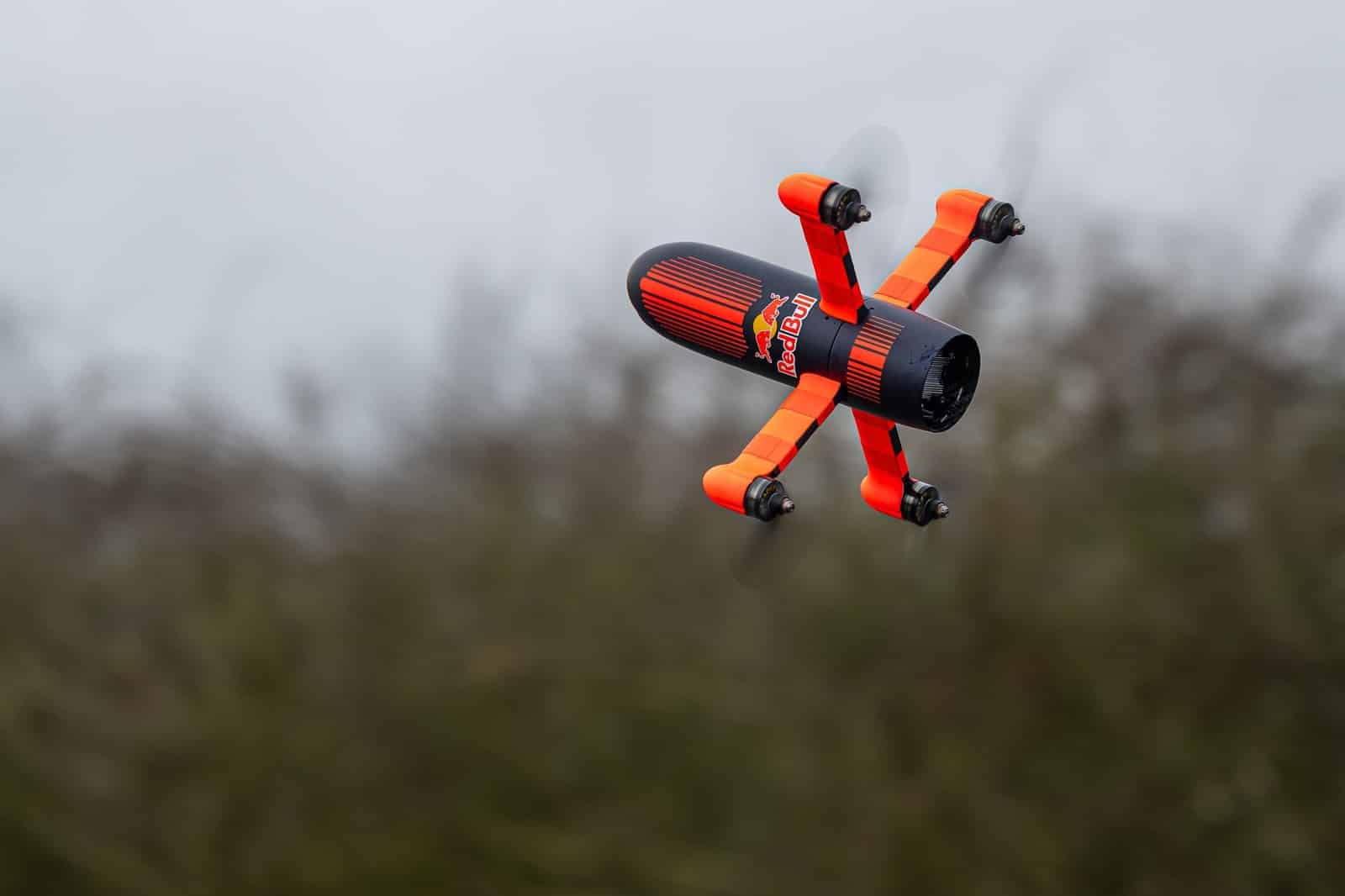 An orange and black FPV racing drone streaks past a blurred hedgerow.