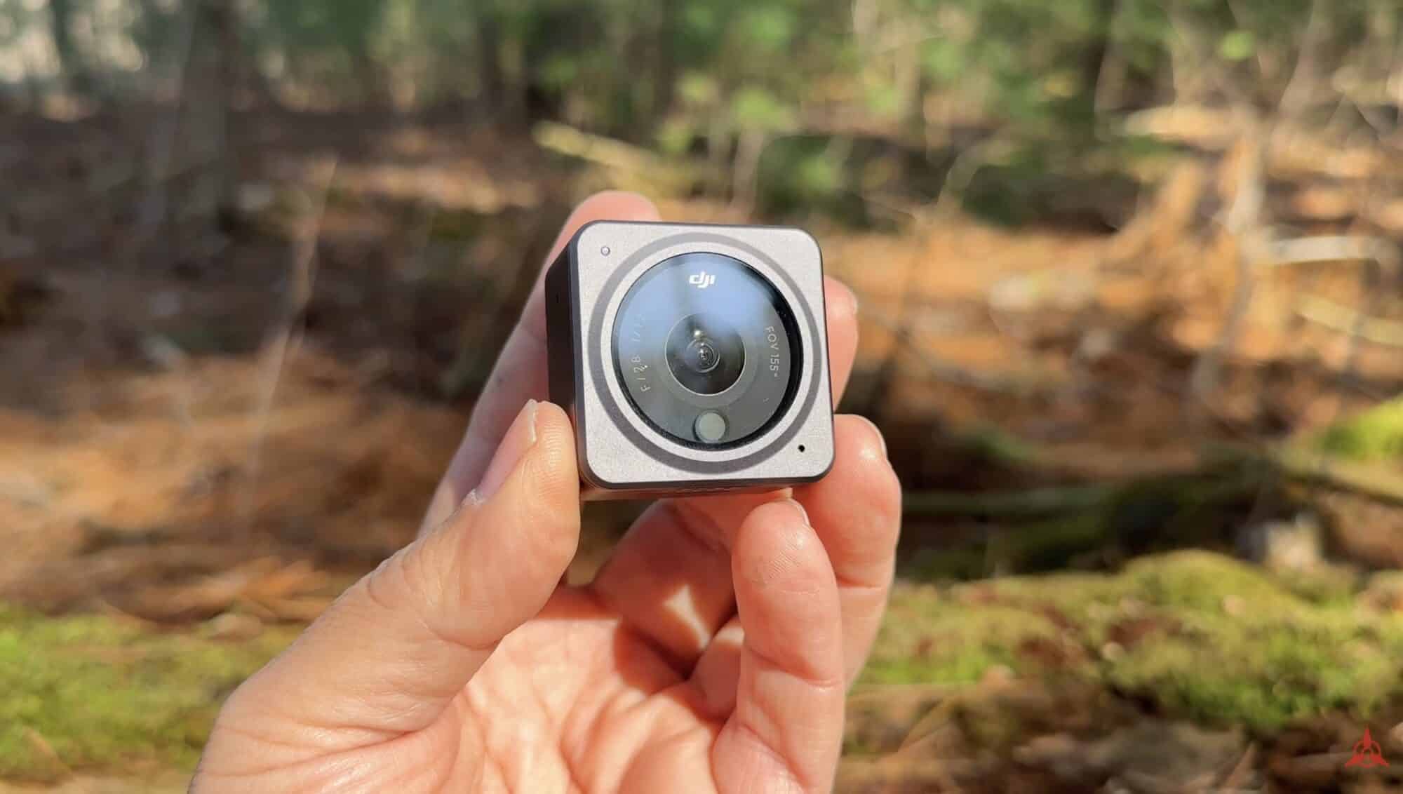 A hand holds the compact DJI Action 2 camera module outdoors in woodland.
