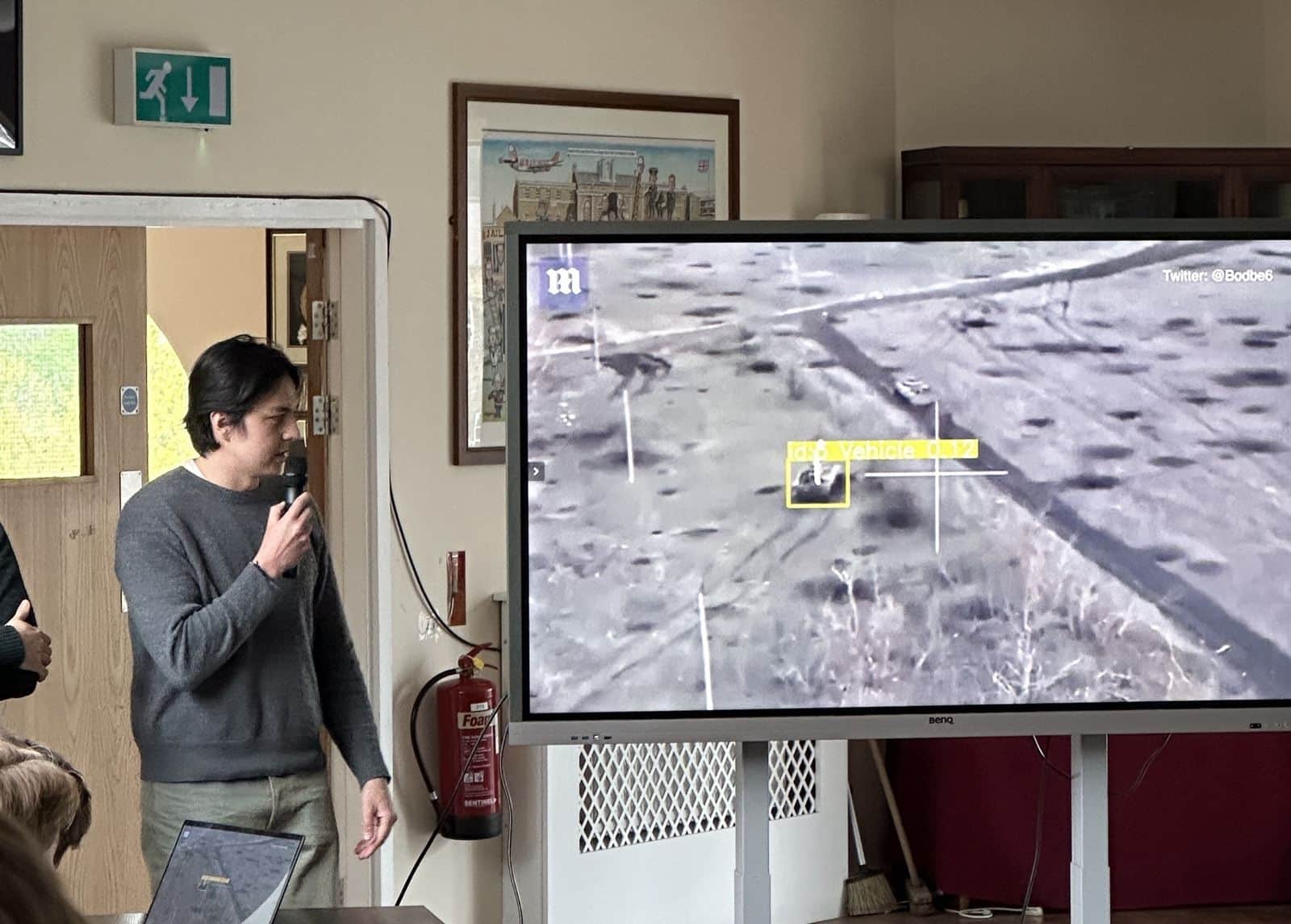 Hackathon presenter beside a screen showing vehicle detection boxes on aerial footage.