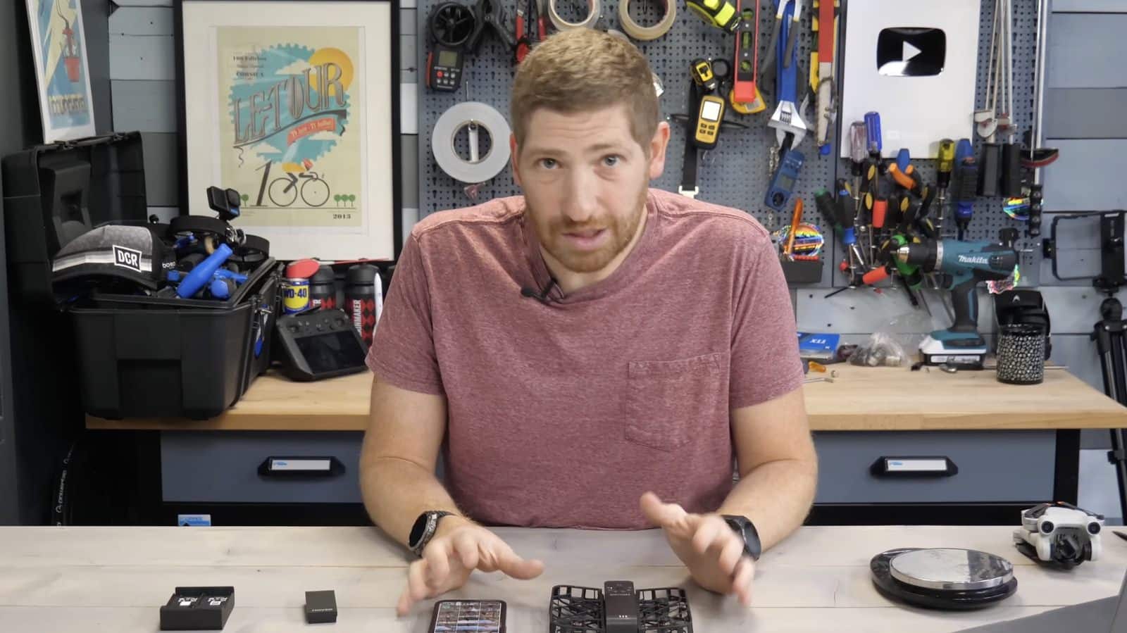 Reviewer gestures at a workbench with HoverAir X1 batteries and accessories spread out.