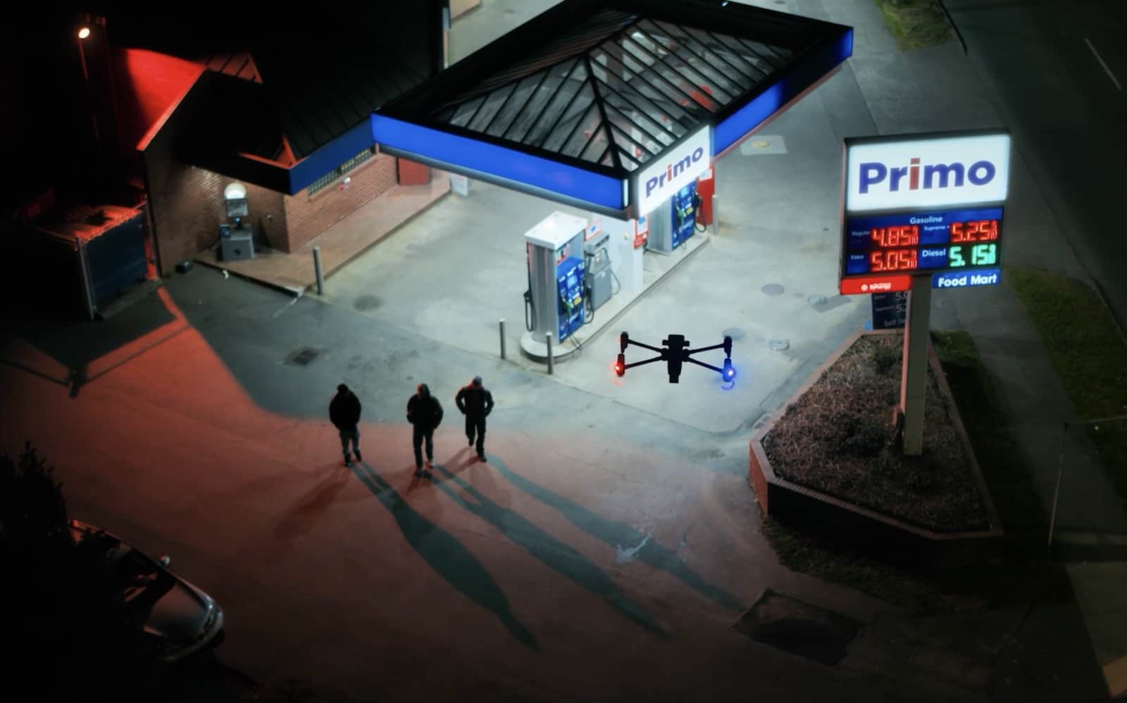 Night aerial of a drone hovering over three people at a lit gas station.