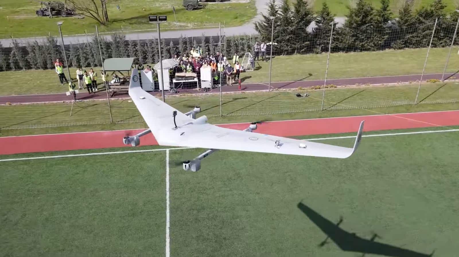 White flying-wing VTOL drone lifting off a sports pitch as observers in hi-vis vests watch.