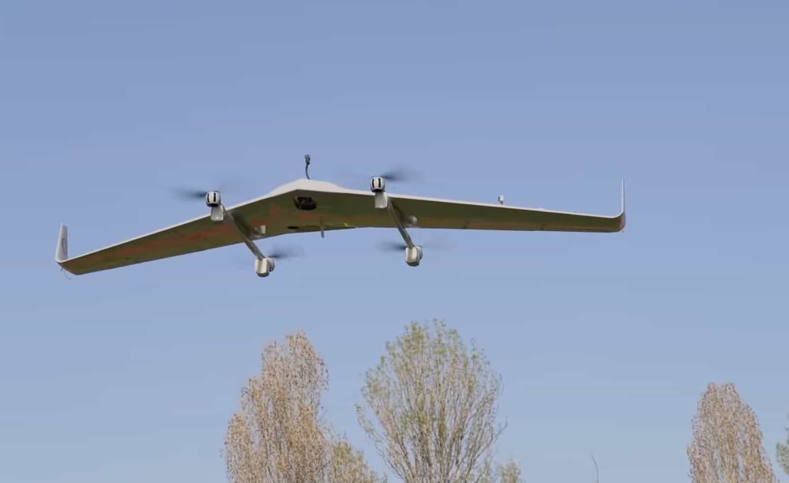 Underside of the flying wing cruising above bare trees with its lift rotors idle.