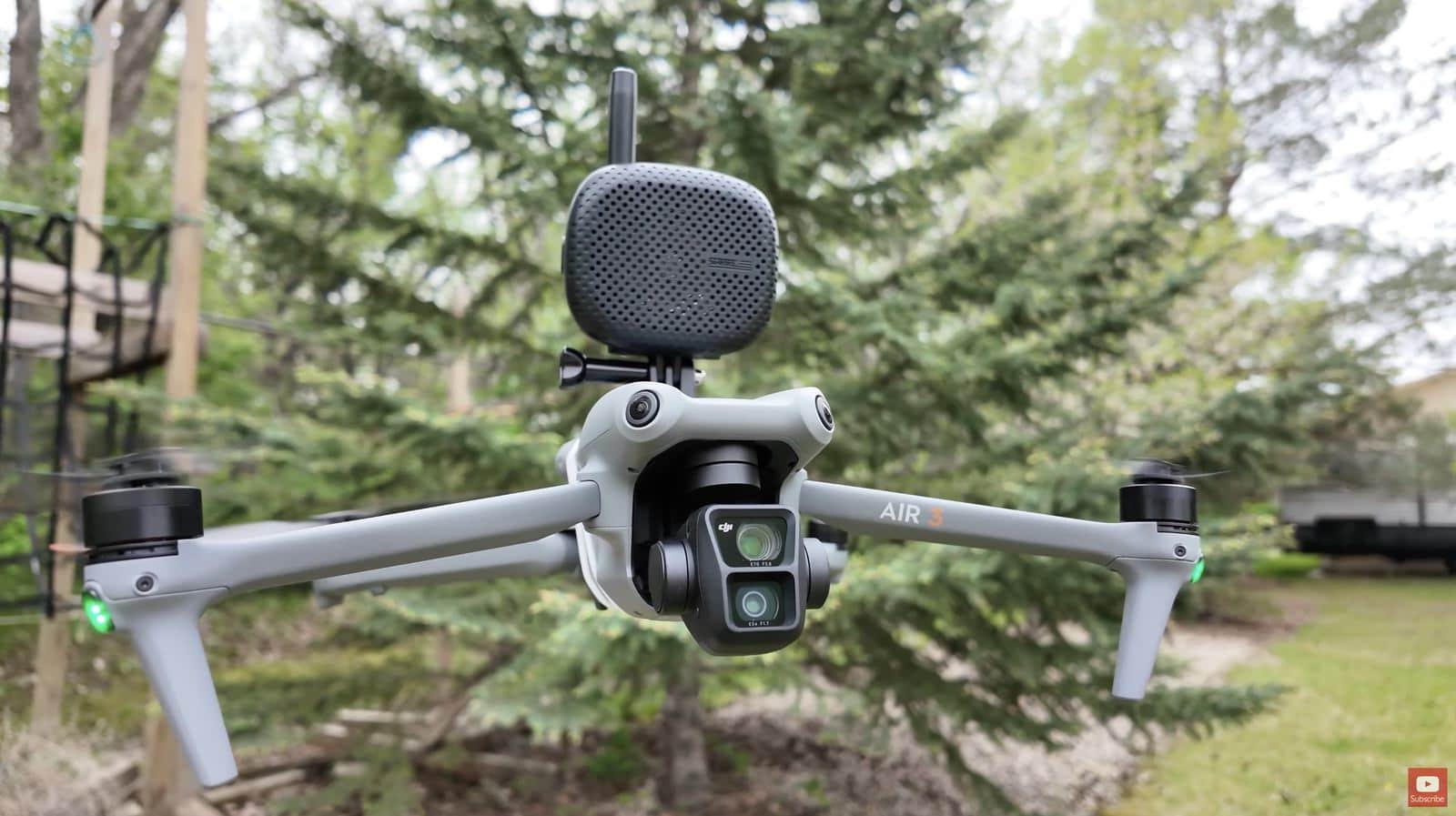 DJI Air 3 hovering beside spruce trees with the speaker strapped above its arms.
