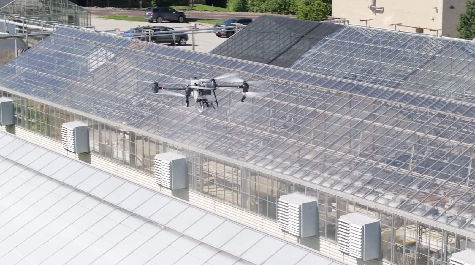 White agricultural spray drone flying low along a glass greenhouse roof.