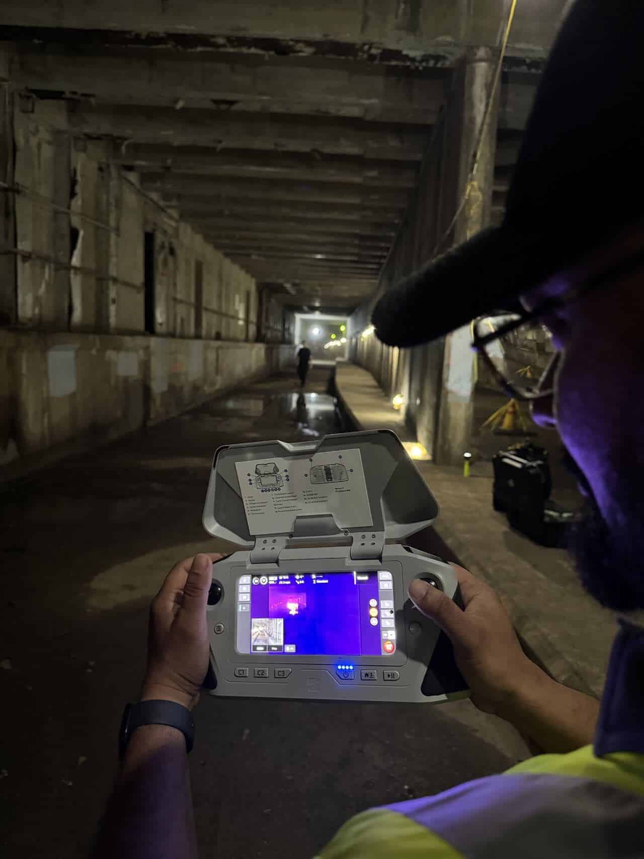An operator flies in a dark subway tunnel with a thermal feed on the controller.