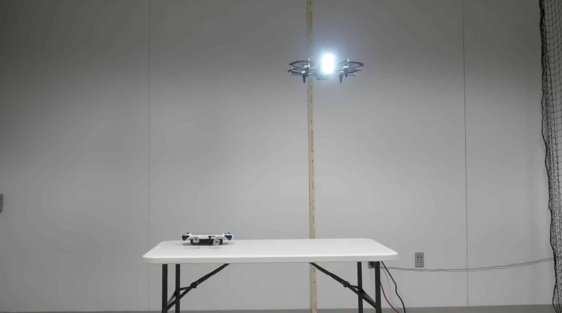 Small drone with a spotlight hovering above a table in a netted test room.