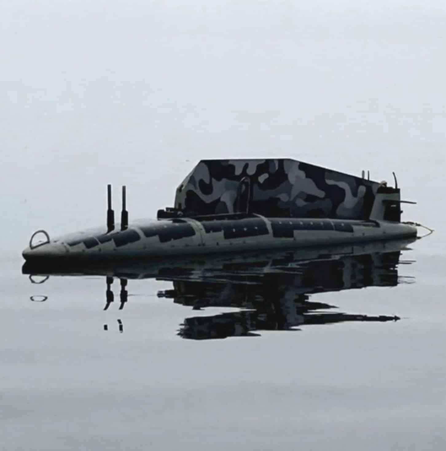 A camouflaged low-profile unmanned vessel sits nearly submerged in calm water.