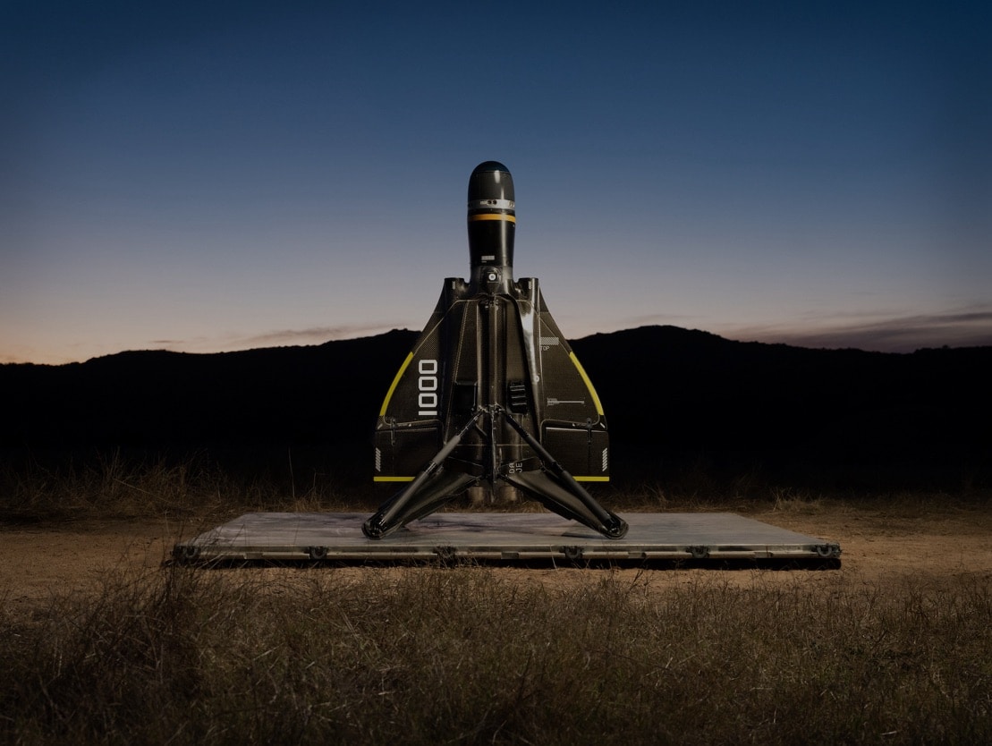 Anduril's Roadrunner munition stands on its launcher at dusk in open scrubland.