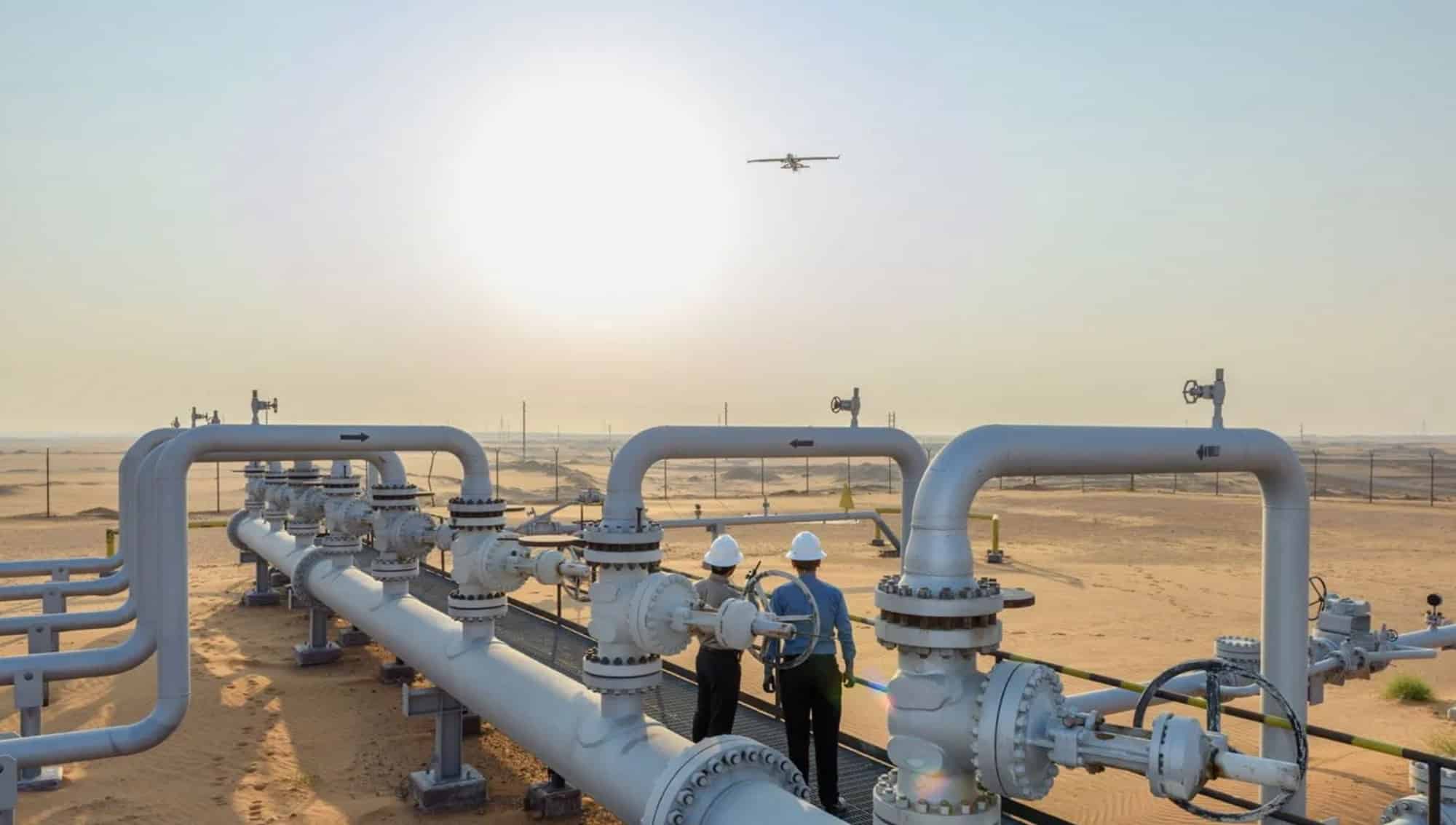 Aramco workers in hard hats at desert wellhead piping as a drone passes overhead.
