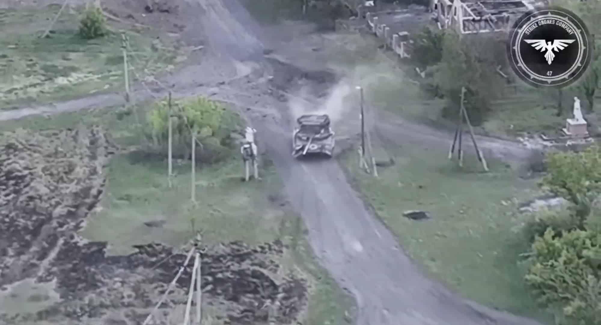 Surveillance drone view of a tank driving a dirt track past ruined village buildings.