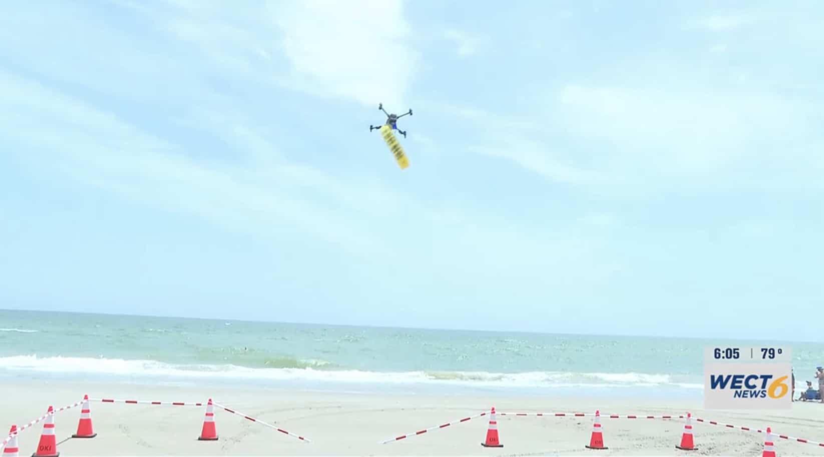 Drone flying over the beach carrying a yellow rescue tube above cone-marked sand.