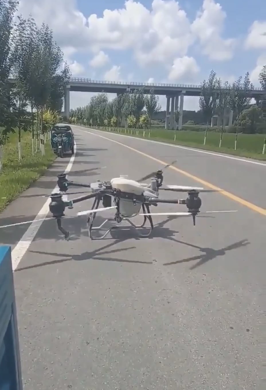 DJI Agras spray drone hovering low over a rural road with a viaduct behind.
