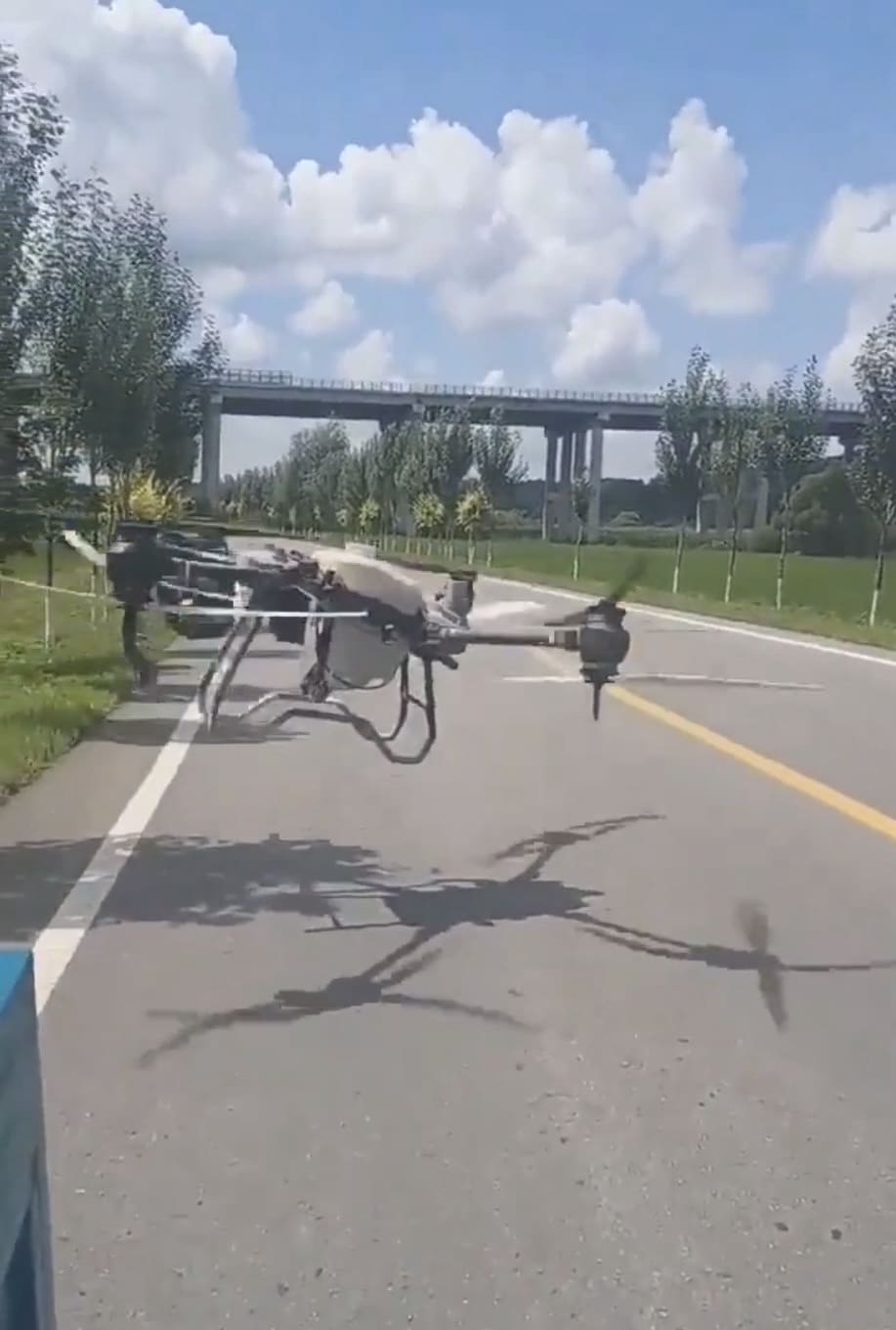 The Agras drone edging forward along the road, its rotor shadow on the asphalt.