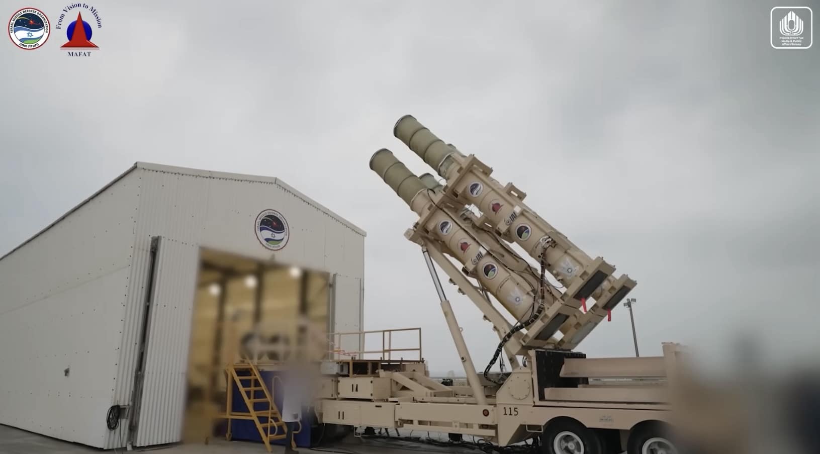 Ground-level view of the launcher tilting four interceptor canisters skyward beside a hangar.