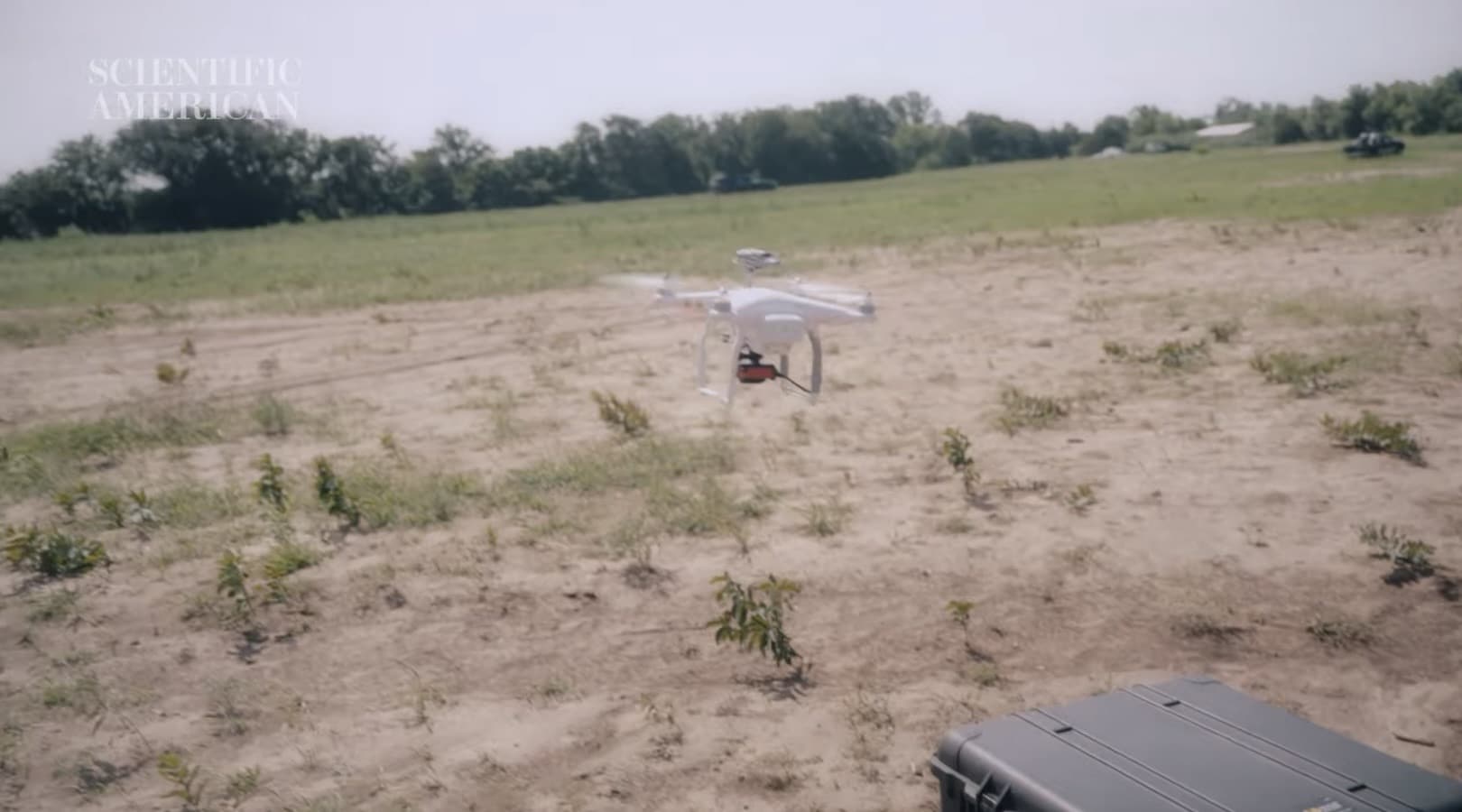 White quadcopter hovers low over a sandy test field beside a hard equipment case.