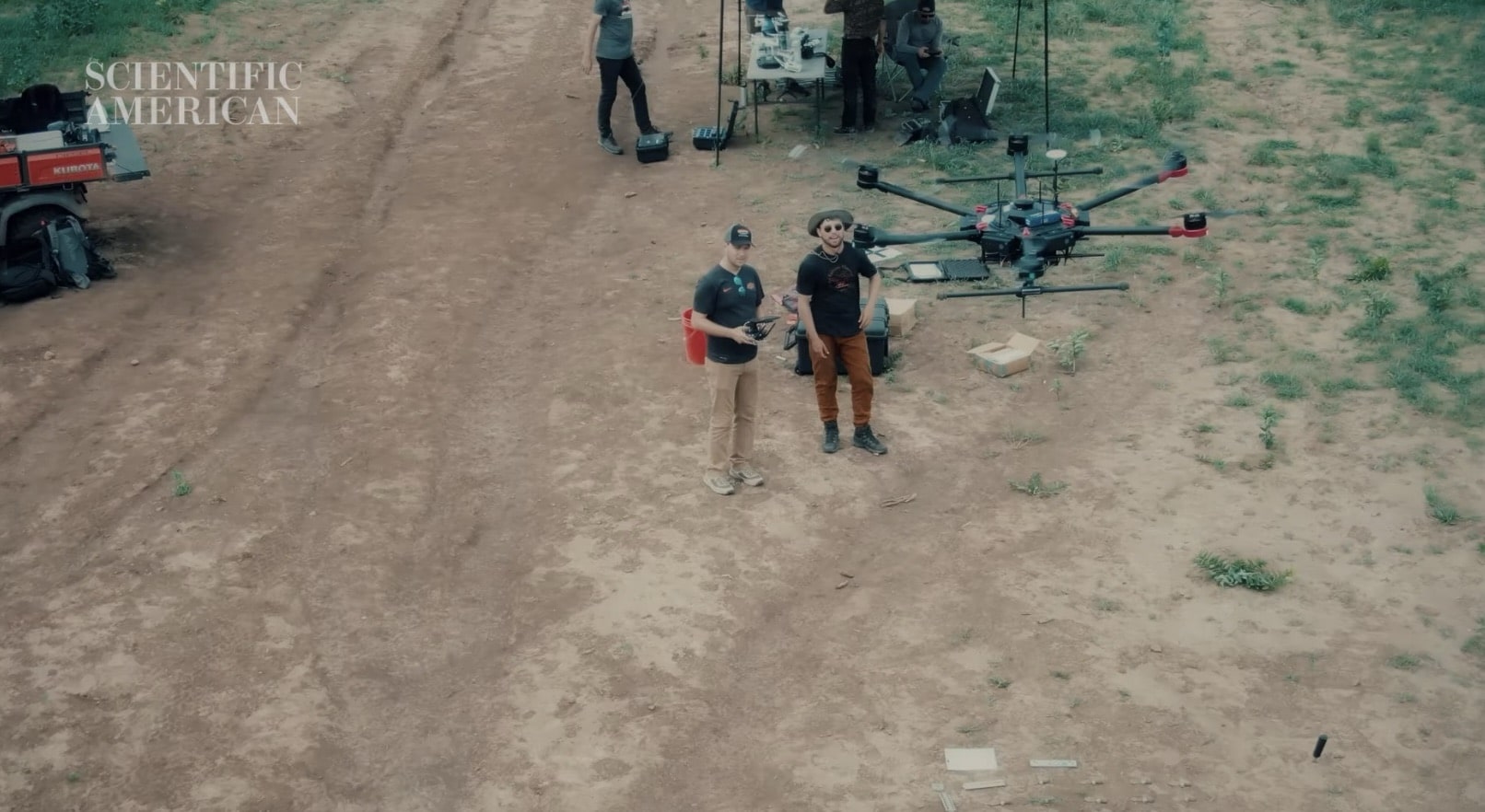 Two operators stand beside a large hexacopter on a dirt track at a test site.