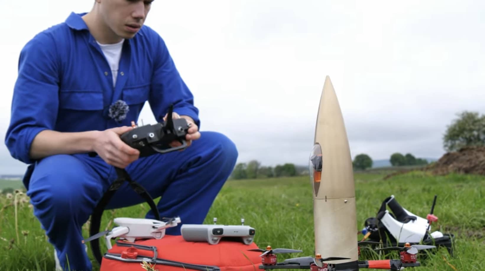 Builder in blue coveralls kneeling with the gold rocket shell and quadcopter frames.