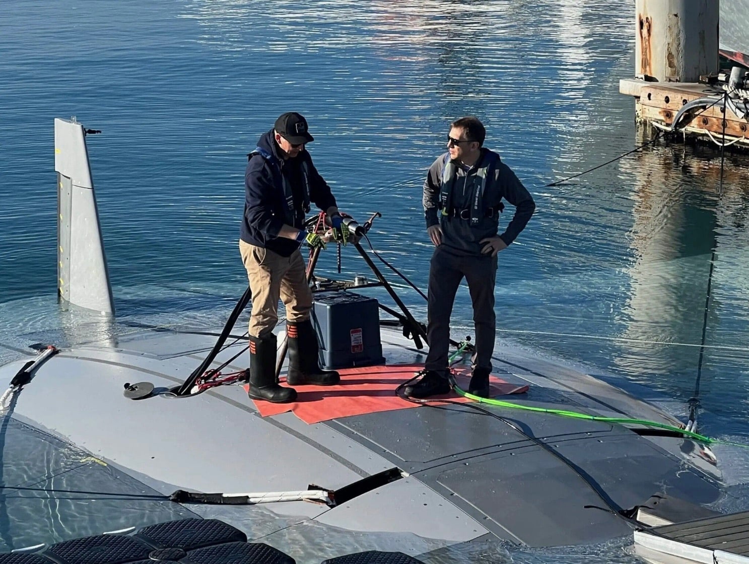Two technicians standing on the surfaced Manta Ray underwater drone beside a dock.