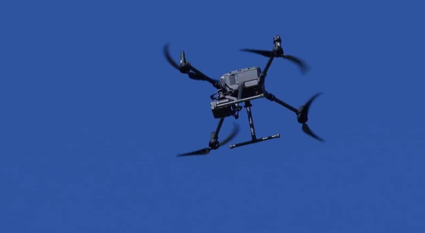 Dark quadcopter with a gimbal camera hovering against a clear blue sky.