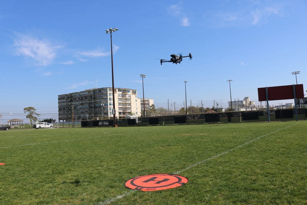 Larger quadcopter hovering over the same field beside an orange landing pad.