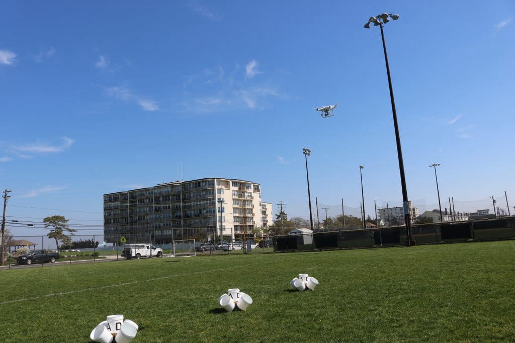 Small white training drone flying above a grass sports field near an apartment block.