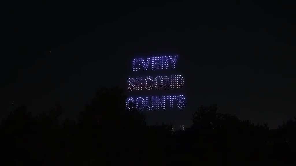 Drone light show spelling EVERY SECOND COUNTS in the night sky above a treeline.