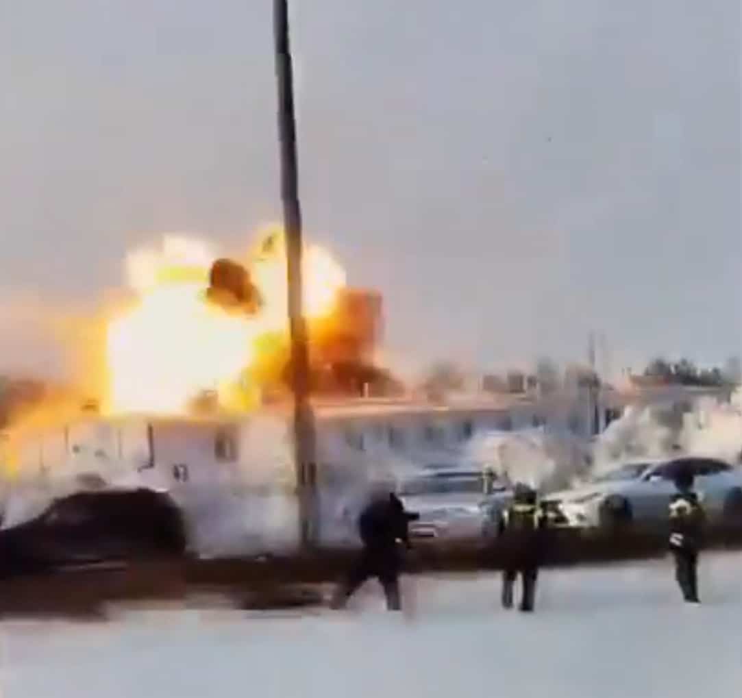 Fireball erupting behind a low building on a snowy street as bystanders run.