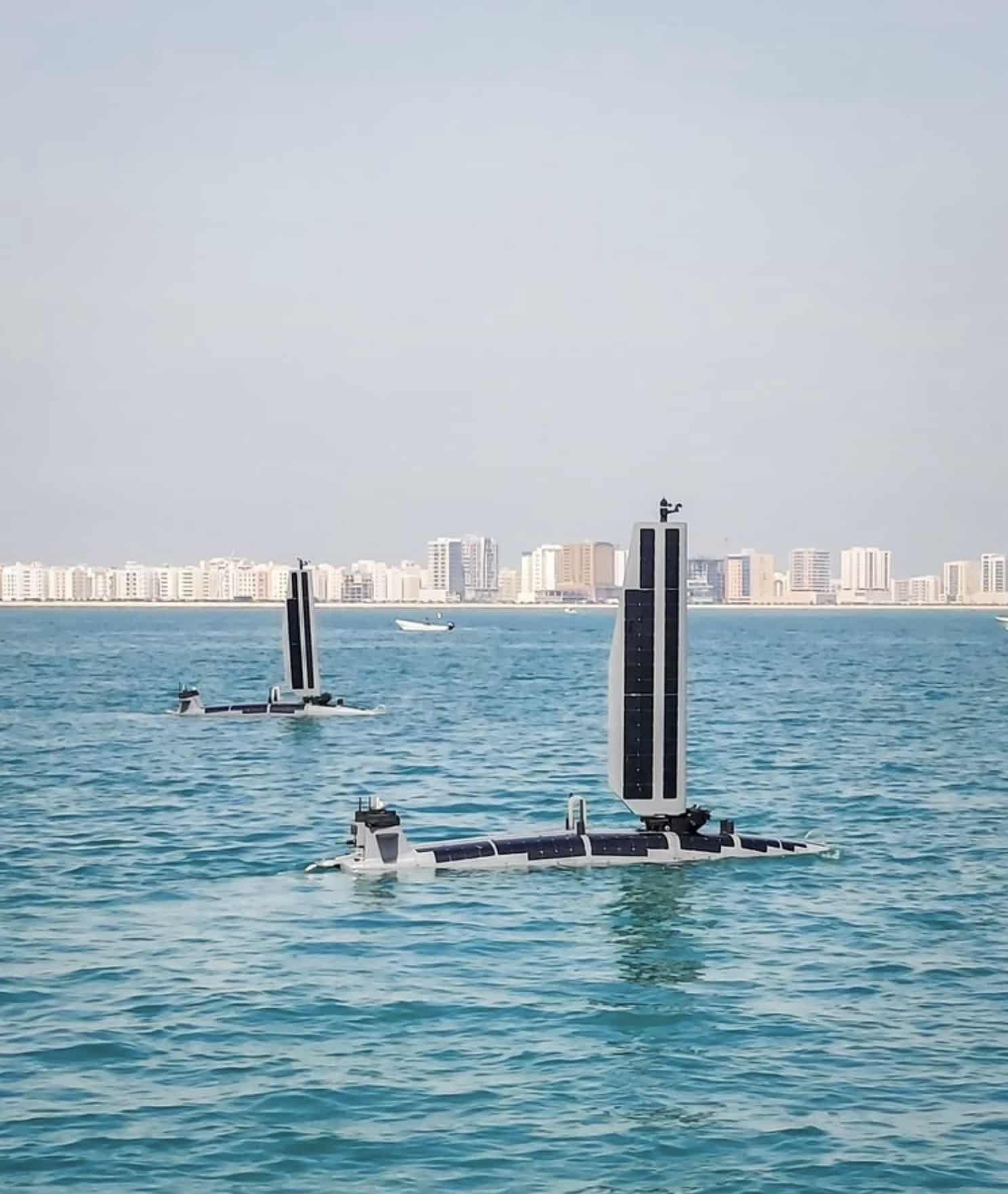 Two solar-powered unmanned surface vessels transit calm water off a city skyline.