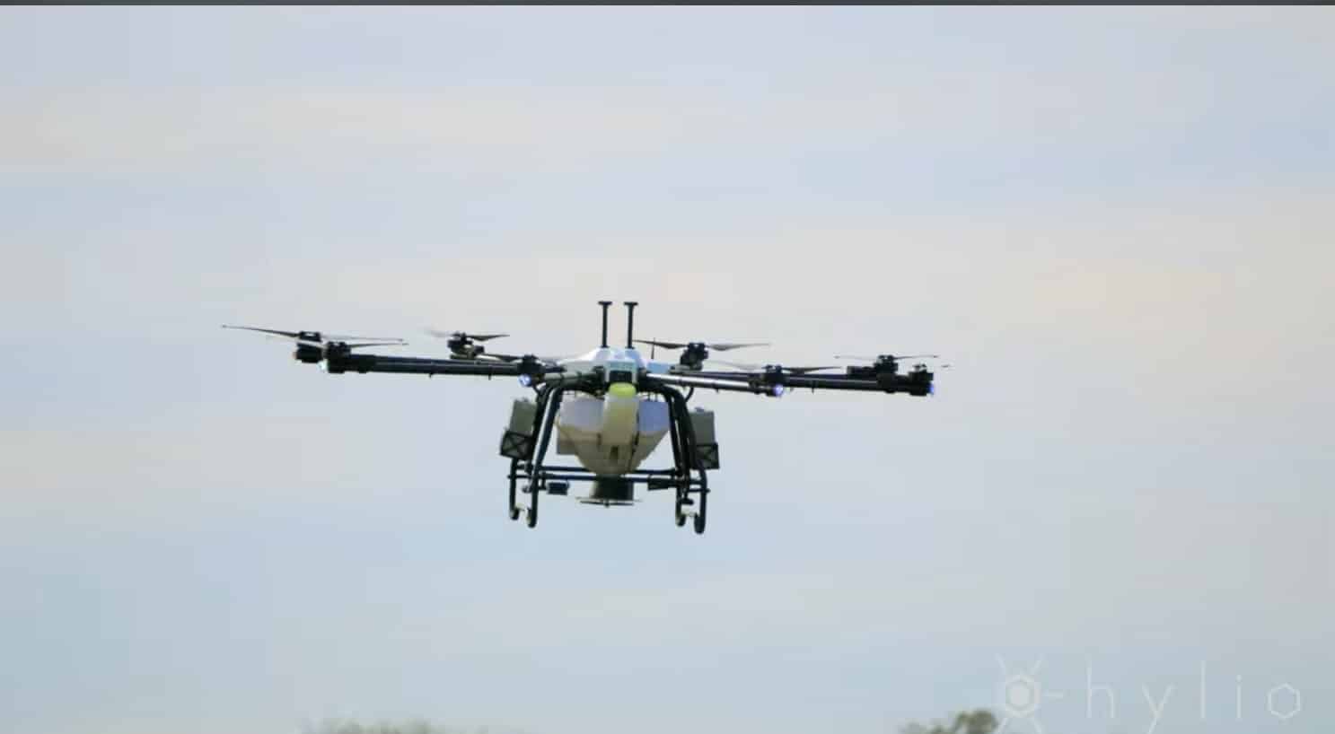 Hylio agricultural spray drone hovering with its tank and spray booms against pale sky.