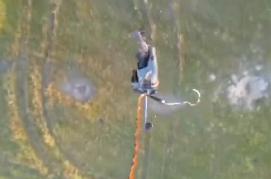 Overhead frame of a quadcopter lowering an orange line and hook over grassland.