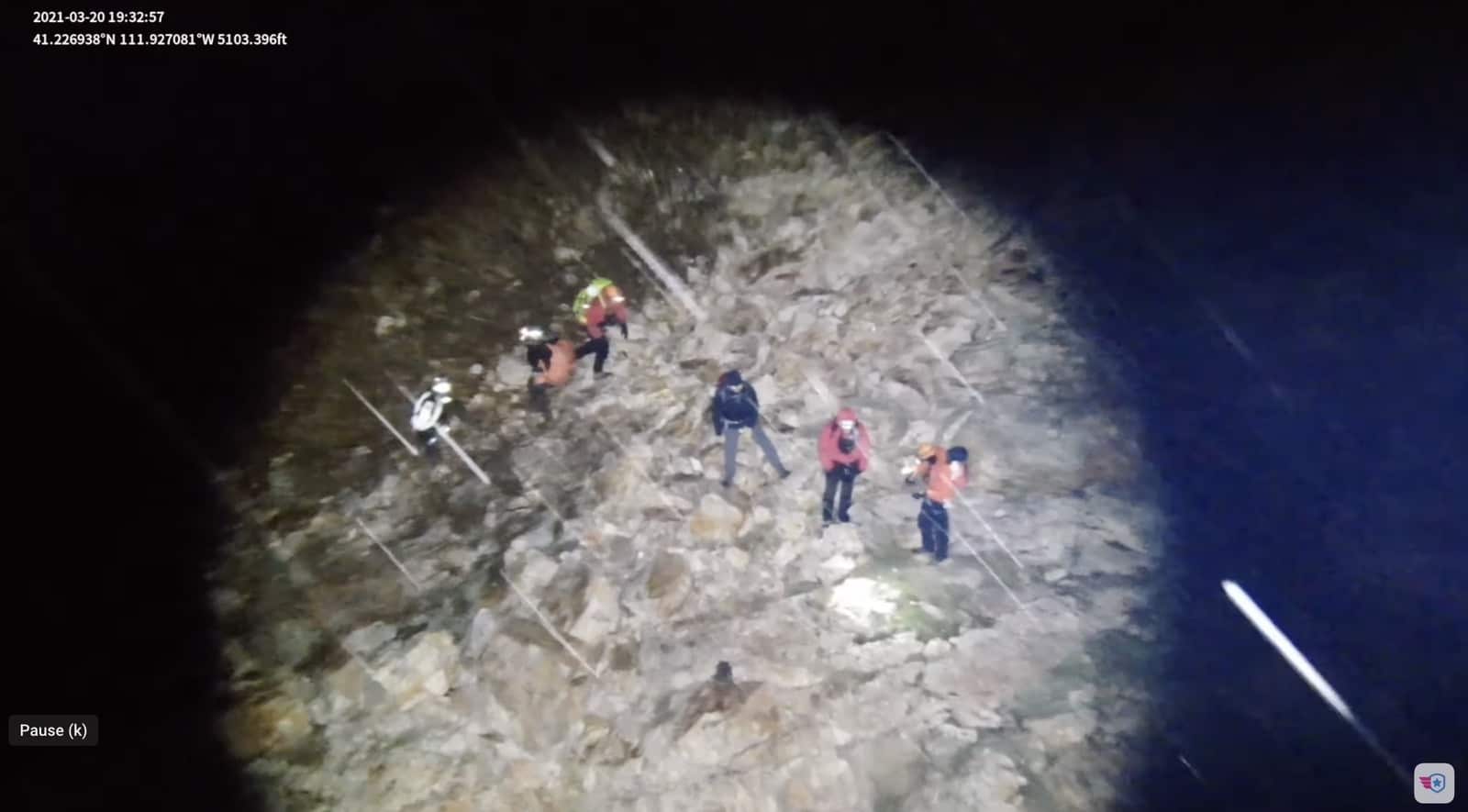 Night drone spotlight view of rescuers gathered around a subject on a rocky slope.