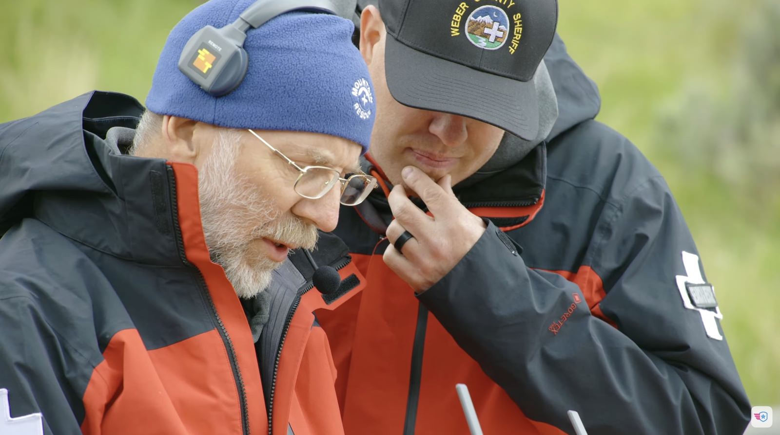 Two rescuers in red jackets conferring, one speaking into a handheld radio.