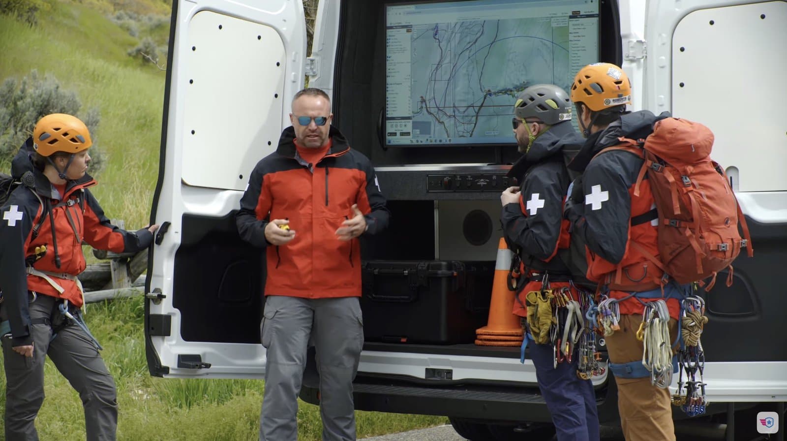 Team leader briefing helmeted rescuers with packs beside the open command van.