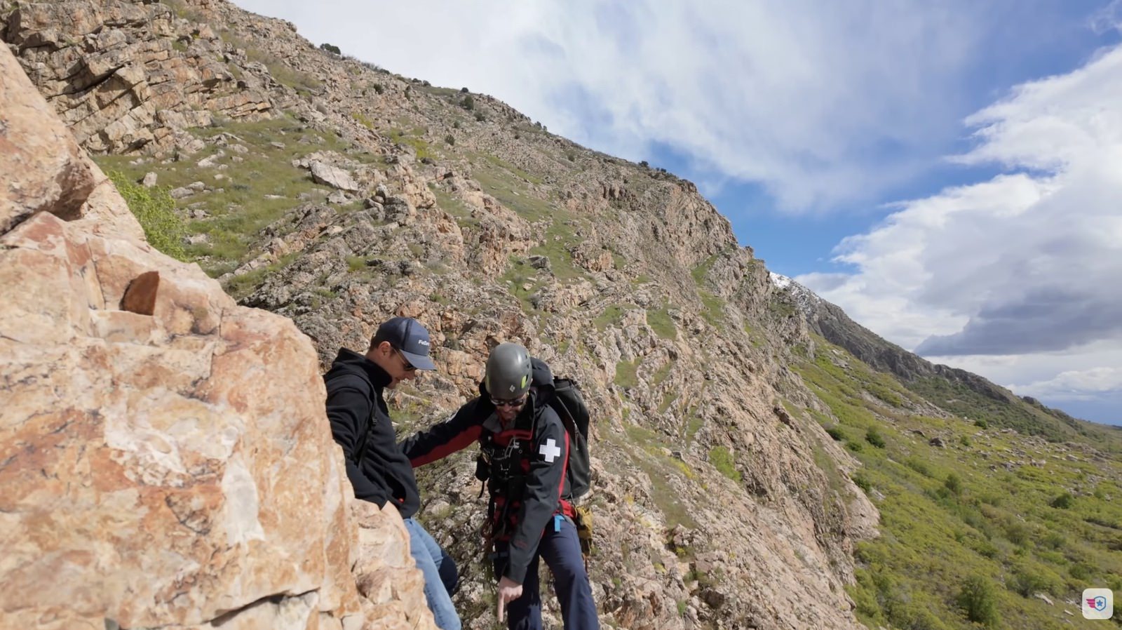 Rescuer in a helmet reaching down to a hiker on a steep rocky slope.