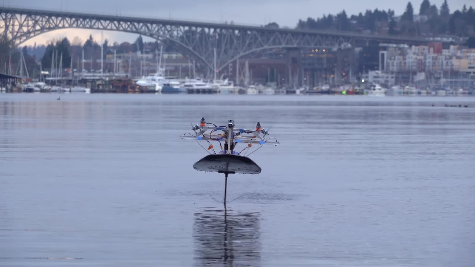 Drone-powered hydrofoil board flying above flat water in front of a marina and bridge.