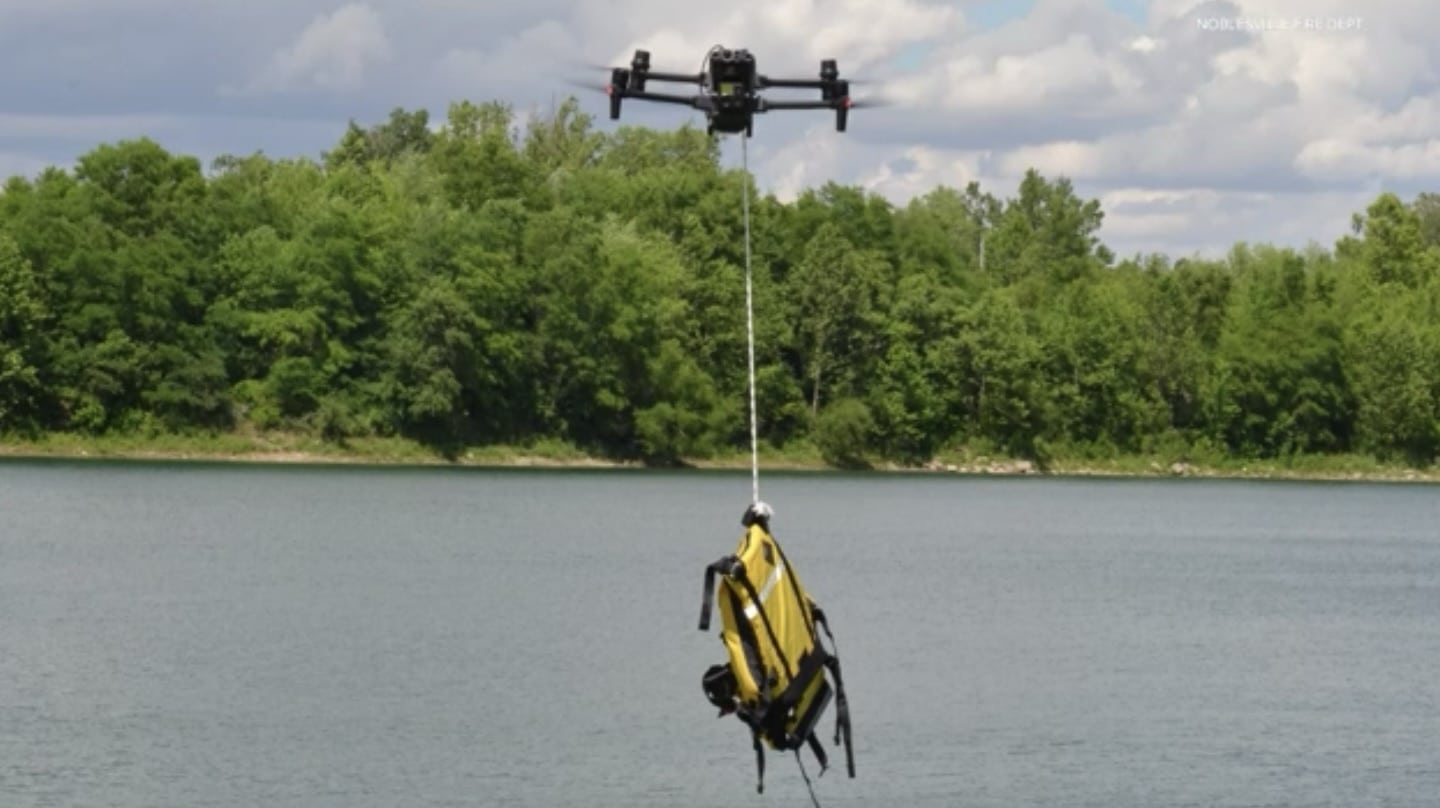 Quadcopter lowers a yellow flotation rescue device on a line above a lake.