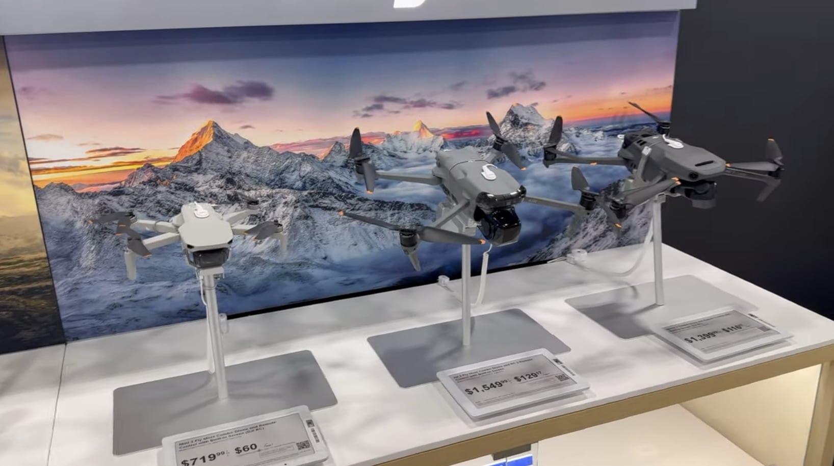 Three DJI drones on Best Buy display stands in front of a mountain backdrop.
