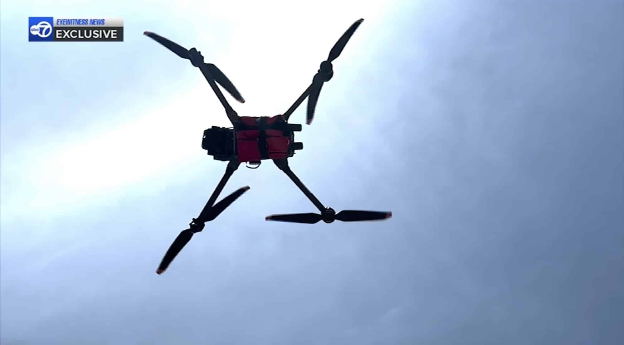 Drone silhouetted from below against a bright sky, red payload visible under the frame.