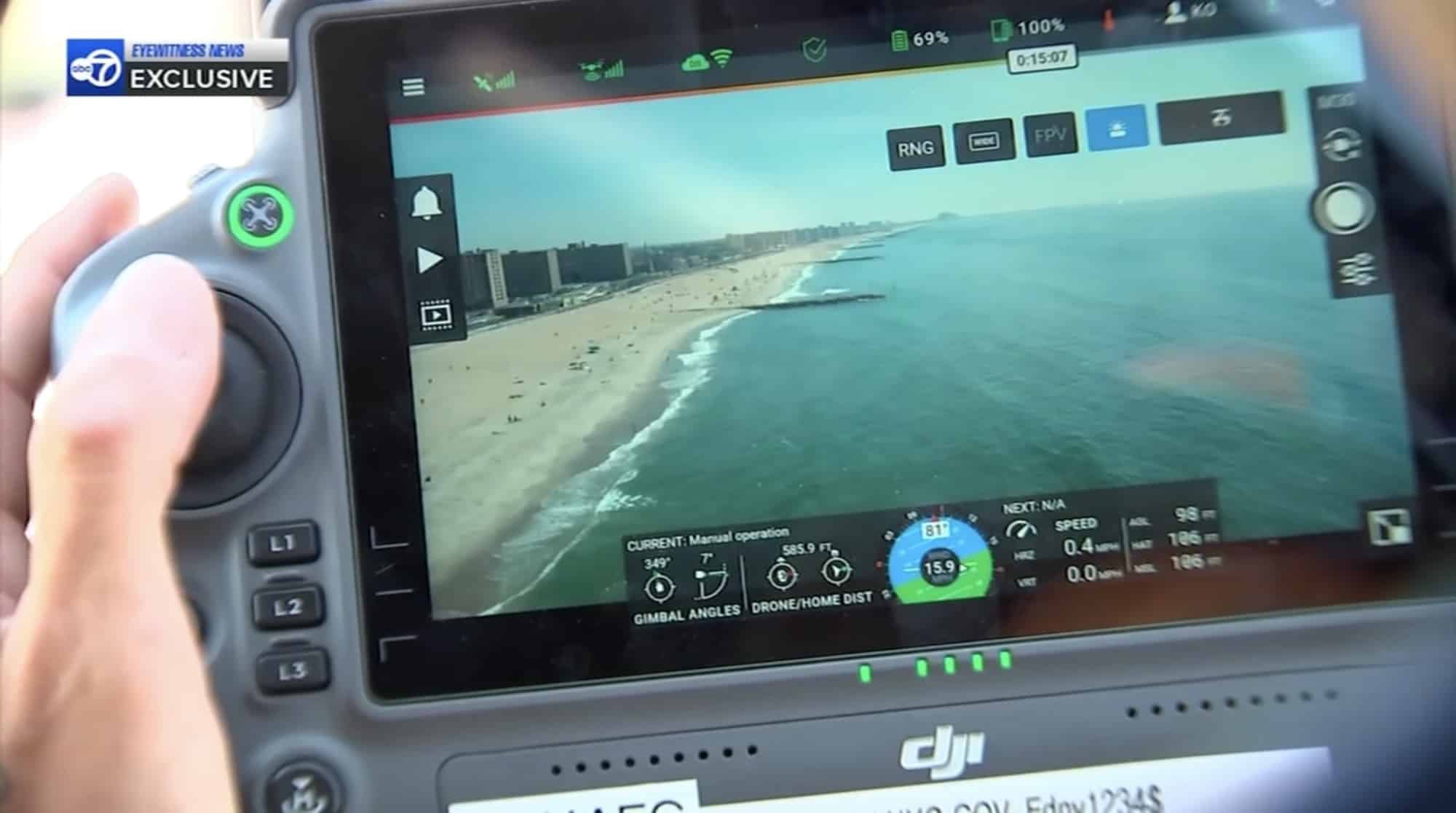 Hands on a DJI controller whose screen shows a live aerial view of the shoreline.