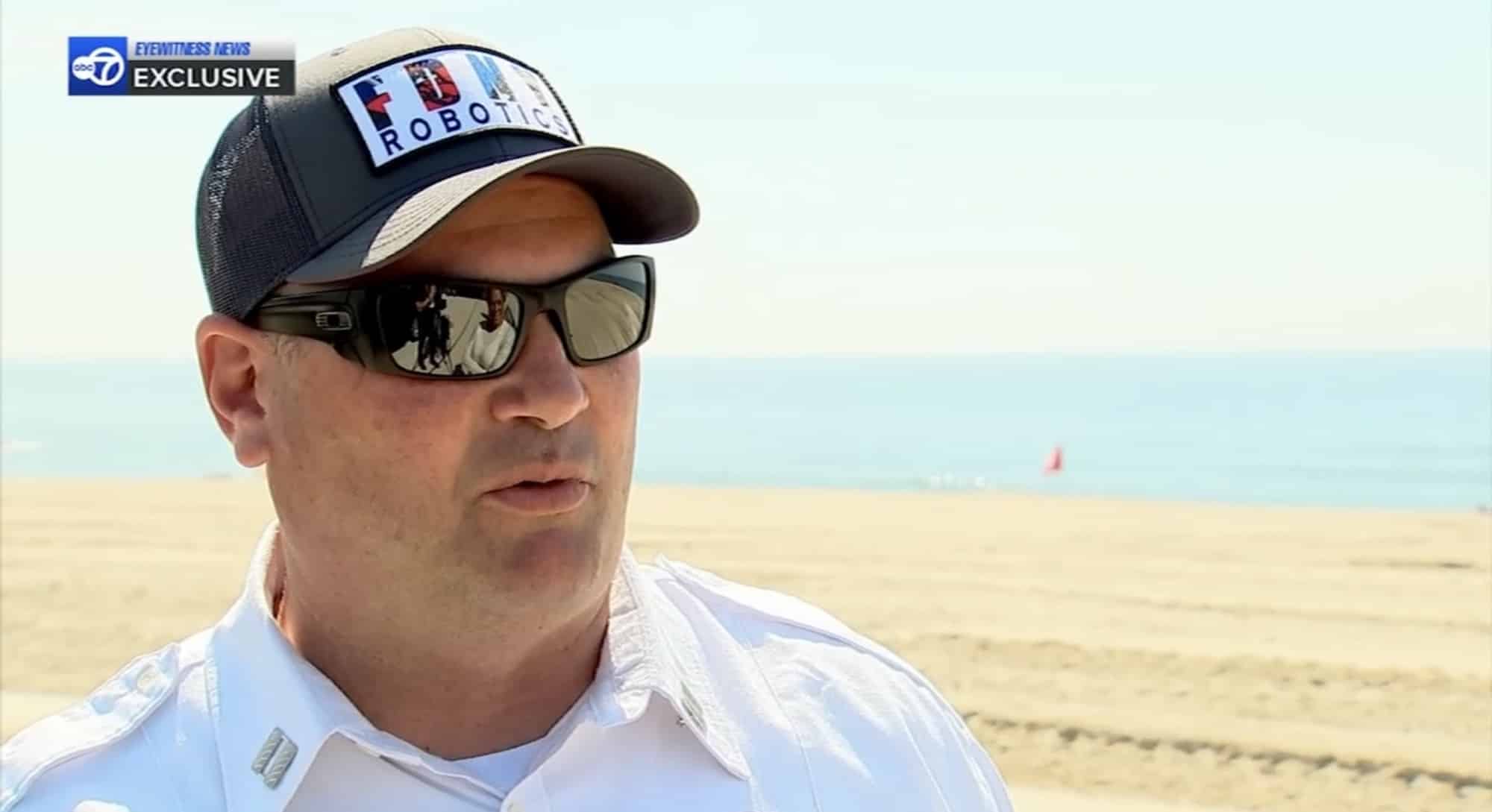 Officer in a white uniform shirt and Robotics ball cap interviewed on the beach.