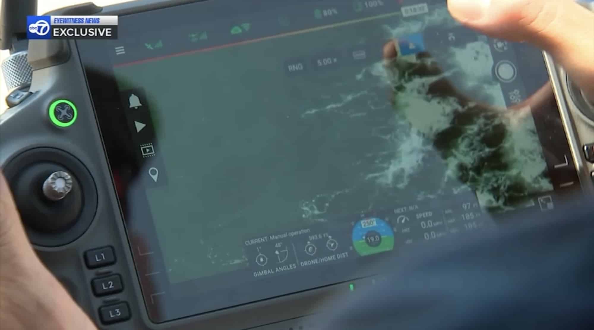 Fingertip on a controller screen showing a drone view of rocks in the surf.