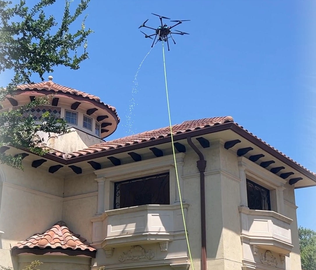 Tethered wash drone spraying a tile-roofed building from above with its supply hose trailing.